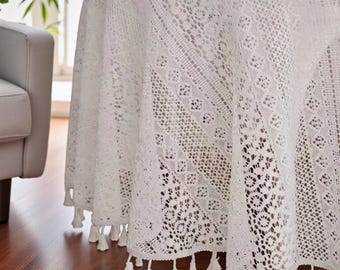 May include: A white, round lace tablecloth with a detailed openwork pattern. The tablecloth has a scalloped edge with small tassels. The table covering is draped over a round table, with a glimpse of a newspaper and a white object on top.