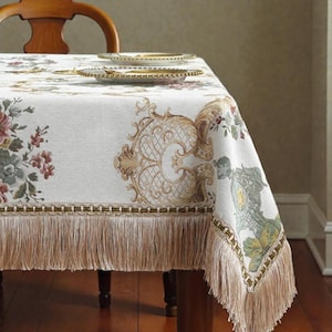 May include: Cream-colored tablecloth with a floral and ornate design. The tablecloth features a gold fringe along the bottom edge and is draped over a wooden table. Two plates with gold rims are stacked on the table.
