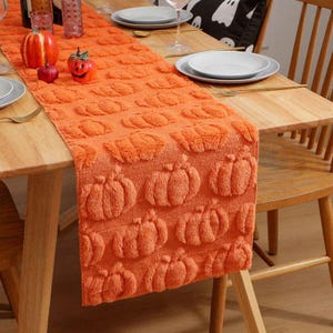 May include: An orange table runner with a repeating pumpkin pattern. The runner is made of a textured fabric and is placed on a wooden table. Decorative pumpkins and plates are also on the table, creating a festive fall theme.