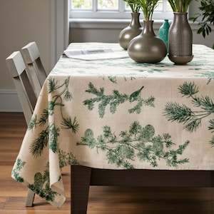 以下が含まれることがあります： クリーム色の背景に緑色の松の枝と鳥の模様が描かれた長方形のテーブルクロス。 テーブルクロスは濃い茶色の木製テーブルに掛けられています。 3つの装飾的な花瓶がテーブルの上にあります。