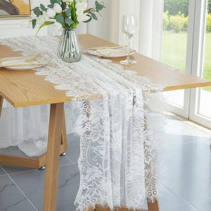 Può includere: Un tavolo da pranzo in legno chiaro è adornato da un runner in pizzo bianco. Il runner presenta intricati motivi floreali lungo i bordi. Un vaso di fiori bianchi e un bicchiere da vino sono sul tavolo, creando un'atmosfera elegante.