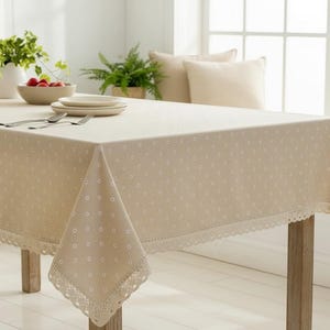 May include: Cream-coloured tablecloth featuring a small polka dot design and a lace border. The rectangular cloth drapes over a wooden table, creating an elegant setting. The table is set with plates, cutlery, and a bowl of fruit.