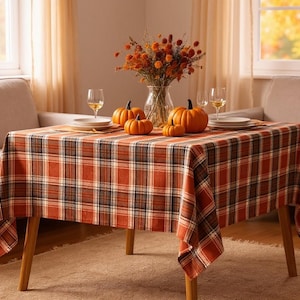 May include: A square dining table covered with an orange, brown, and white plaid tablecloth. The table is set with a clear glass vase containing dried orange flowers, three orange pumpkins, two white plates, and two clear wine glasses.