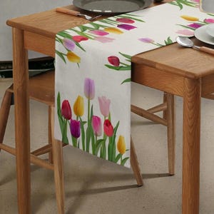 Puede incluir: Camino de mesa rectangular con un diseño floral de tulipanes. El camino de mesa presenta un fondo blanco con tulipanes coloridos en tonos de rojo, rosa, morado y amarillo. También se ven hojas y tallos verdes.