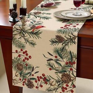 May include: Beige table runner with a festive winter design. The runner features illustrations of pinecones, red berries, and evergreen branches. A red bird is also depicted. The runner is made of a woven fabric.