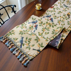 Op de afbeelding: Beige tafelloper met een vogel- en bloemenpatroon. De loper toont blauwe vogels op takken met groene bladeren en gele bloemen. De randen zijn afgewerkt met kwasten in blauwe en bruine tinten. Geschikt voor eettafels.