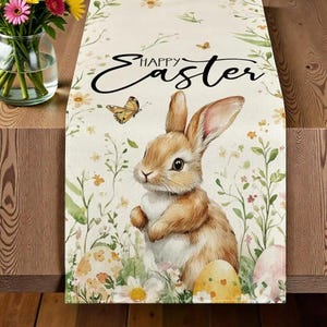 May include: Easter-themed table runner featuring a brown bunny, decorated Easter eggs, and floral accents. The words "Happy Easter" are written in a cursive font. The runner is made of a light-colored fabric.