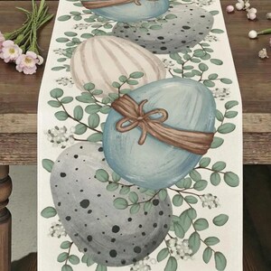 Può includere: Runner da tavolo a tema pasquale con uova dipinte in colori pastello, tra cui blu, rosa e grigio, decorate con nastri marroni e circondate da foglie verdi e fiori bianchi. Per la decorazione.