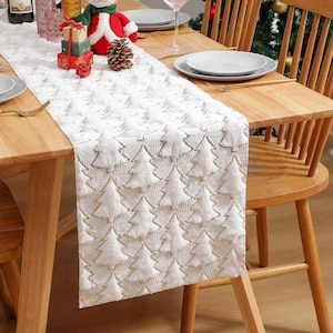 May include: White table runner with a repeating pattern of Christmas trees in a textured design. The runner is placed on a wooden table, with plates, silverware, and decorative items visible. The runner adds a festive touch to the dining setting.