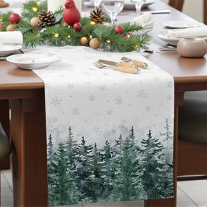 Könnte beinhalten: Weißer Tischläufer mit einer Winterwaldszene am unteren Rand und Schneeflocken darüber. Der Wald ist in Grün- und Grautönen dargestellt. Der Läufer liegt auf einem Holztisch, der für ein Essen gedeckt ist.