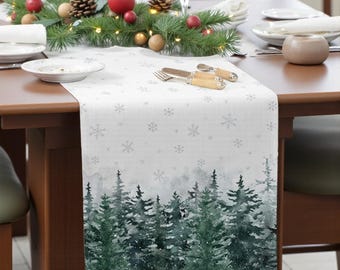 Puede incluir: Camino de mesa blanco con una escena de bosque invernal en la parte inferior y copos de nieve dispersos en la parte superior. El bosque está representado en tonos verdes y grises. El camino de mesa está sobre una mesa de madera.