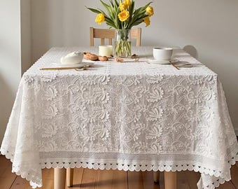 May include: A white lace tablecloth with a floral pattern covers a wooden table. The tablecloth has a scalloped edge. A vase of yellow flowers, a teacup, and a plate are on the table.