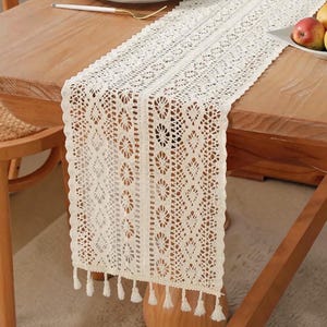 Puede incluir: Camino de mesa de encaje crema de tejido abierto con un patrón floral y flecos de borlas. El camino de mesa se muestra sobre una mesa de madera, agregando un toque de elegancia. El diseño intrincado es visible.