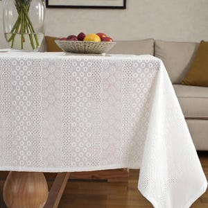 Pode incluir: Uma toalha de mesa branca com um padrão floral e de pontos detalhado cobre uma mesa de madeira. Um vaso de vidro com rosas cor-de-rosa e uma tigela de frutas estão sobre a mesa. A toalha de mesa cai pela borda.