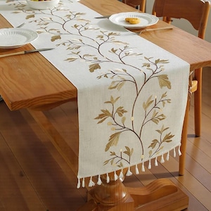May include: Cream-coloured embroidered table runner with a botanical design. The runner features a repeating pattern of brown stems and gold-coloured leaves. The bottom edge is finished with white tassels. The runner is displayed on a wooden table.