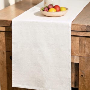 May include: A white textured table runner on a wooden surface. A bowl of fruit, including strawberries, lemons, and an apple, sits on the runner. The runner's texture adds visual interest to the neutral colour.