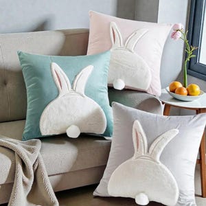 Könnte beinhalten: Vier quadratische Zierkissen in Pastellfarben, jedes mit einem weißen Hasenmuster. Die Ohren und der flauschige Schwanz des Hasen sind deutlich sichtbar. Die Kissen liegen auf einem grauen Sofa und schaffen eine gemütliche Osteratmosphäre.