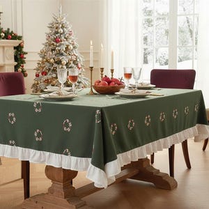 May include: A rectangular dark green tablecloth featuring a repeating pattern of small embroidered wreaths and a white ruffled border along the bottom edge. The fabric drapes over a wooden table set with plates, glasses, and a bowl of red fruit.