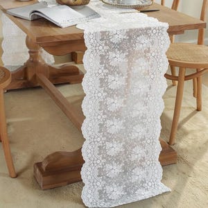 Puede incluir: Un camino de mesa de encaje blanco con un patrón floral, colocado sobre una mesa de madera. El camino de mesa tiene un borde festoneado y añade un toque delicado al comedor. La mesa es de madera y tiene un acabado cálido y natural.