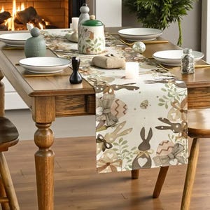 May include: A wooden dining table set for a meal, featuring an Easter-themed table runner with bunny and egg designs. The table is set with white plates, gold-coloured utensils, and decorative ceramic pieces.
