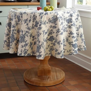 Peut inclure: Une table ronde en bois avec une nappe crème ornée d'un motif floral bleu. La table, avec un piedestal, est installée dans une cuisine. La nappe recouvre la table.