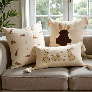 May include: A set of three decorative throw pillows with bunny designs. The pillows are cream-colored with embroidered brown rabbits and tassels. One pillow features a large bunny design.