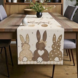 Könnte beinhalten: Beiger Ostern-Tischläufer mit drei braunen Hasen-Silhouetten mit Herz- und Tupfenmustern. Jeder Hase hat eine Schleife und einen flauschigen Schwanz. Der Läufer ist mit Osterei-Designs verziert.