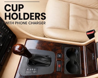 Könnte beinhalten: Ein schwarzer Kunststoff-Getränkehalter mit integriertem Telefonladegerät für die Mittelkonsole eines BMW E38. Das Bauteil verfügt über zwei runde Öffnungen und befindet sich neben einer hölzernen Schaltkulisse mit schwarzem Lederschaltknauf.