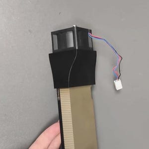 May include: A black electronic component with a rectangular top and a beige base. The component has wires in blue, red, and white. The item is held by a hand.