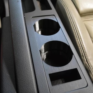 May include: Black car console featuring two circular cup holders and a rectangular slot. The console is constructed from a textured, dark material and is positioned near a beige leather car seat. A gear shift lever is visible.