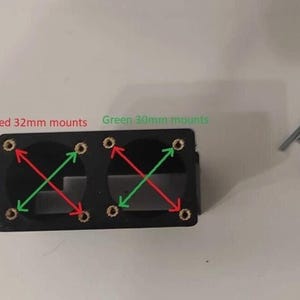 Puede incluir: Un soporte rectangular negro con dos aberturas circulares, con flechas direccionales rojas y verdes que indican soportes de 32 mm y 30 mm. Varios tornillos plateados están dispersos cerca.