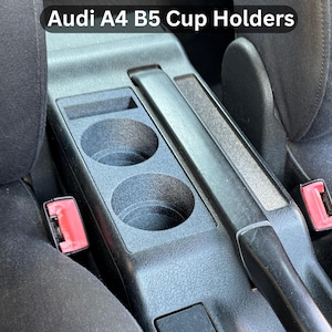 Peut inclure: Porte-gobelets en plastique noir pour une Audi A4 B5. La console comprend deux porte-gobelets circulaires, un espace de rangement rectangulaire et un accoudoir rembourré. Le texte "Audi A4 B5 Cup Holders" est visible.