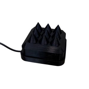 Op de afbeelding: Een zwart, rechthoekig apparaat met een raster van conische spikes. Het apparaat heeft een getextureerd oppervlak en een zwarte draad die zich vanaf de zijkant uitstrekt. De spikes zijn gelijkmatig verdeeld en steken uit het bovenoppervlak.