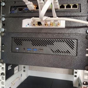 May include: A black server rack mount panel containing a small form factor computer with a front-facing power button, two USB 3.0 ports, and one USB 2.0 port, positioned below a white network switch and a black router with multiple white ethernet cables.