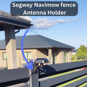 May include: A black antenna holder mounted on a dark gray fence. The text "Segway Navimow fence Antenna Holder" is displayed above the antenna. The background shows a house and a blue sky.
