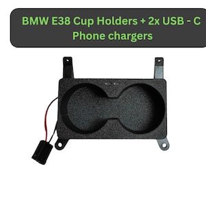 May include: Black cup holder with two circular openings and two USB-C phone chargers. The product has a textured surface and is designed for a BMW E38. The text "BMW E38 Cup Holders + 2x USB-C Phone chargers" is displayed above.