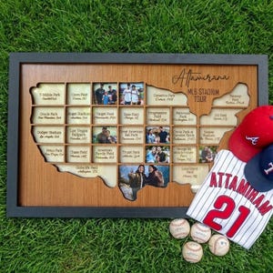 May include: A wooden MLB Stadium Tour map with photo slots, a baseball jersey, two baseball caps, and baseballs on green grass. The map is in the shape of the United States and has the name "Altamirana" written on it.