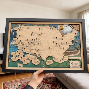 May include: A framed wooden travel map of the United States, featuring a layered design with a natural wood finish. The map includes state outlines, trees, and an eagle illustration. The frame is black, and the map has the text "Travel Map" and the names "Leo & Luna."