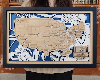 May include: A framed wooden map of the United States with a blue background and a black frame. The map highlights NFL stadiums with small white circles. The design includes football-themed illustrations and the text "MappaePilot NFL Stadium Tour."