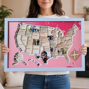May include: A framed wooden map of the United States with photos inserted into each state. The map is set against a pink background and framed in a light blue frame. A wooden compass rose is in the lower right corner.