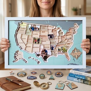 May include: A framed wooden map of the United States with state cutouts, each holding a photograph. The map is set against a light blue background and framed in white. Travel-themed accessories, including pins, a compass, and books, are displayed below.