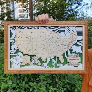 May include: Wooden framed golf-themed artwork featuring a map of the United States with state outlines and golf-related imagery. The artwork includes the text "MappaePilot" and is set against a green and white background.