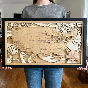 May include: A framed wooden baseball-themed map of the United States, laser-cut with baseballs marking locations. The map includes baseball-related imagery, a baseball glove, and the text "Michael Family". The frame is black.