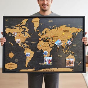 Op de afbeelding: Een wereldkaart met een zwart kader en een kurken overlay. De kaart bevat punaises en foto's die reisbestemmingen markeren. Het frame is zwart en de kaart bevat de tekst "Amy & William Our World Adventures Jan 5 2025."