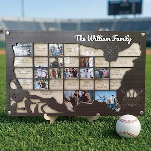 May include: A wooden family photo display in the shape of the USA, with numerous small photo frames. The display is inscribed with "The William Family" and baseball-themed cutouts. A baseball is placed on the grass in front of the display.