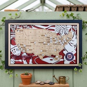 Może przedstawiać: Drewniana mapa Stanów Zjednoczonych z wycięciami o tematyce baseballowej. Mapa zawiera piłki baseballowe oznaczające miasta i logo Washington Nationals. Tło jest czerwono-białe, oprawione w czarno-niebieskie.