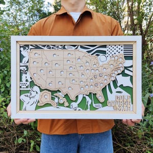 Può includere: Una mappa dei campi da golf in legno incorniciata degli Stati Uniti. La mappa è tagliata al laser con contorni degli stati e illustrazioni a tema golf in tonalità legno naturale e bianco, su uno sfondo verde. La cornice è bianca e il testo "GOLF COURSE MAP" è visibile.