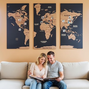 Puede incluir: Decoración mural de mapa del mundo de tres paneles con un fondo azul oscuro y un mapa de corcho. El mapa presenta contornos de países y está adornado con fotos y alfileres. Una pareja mira un álbum de fotos en primer plano.