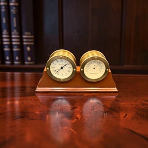 May include: A desk accessory featuring a round Bulova clock with Roman numerals and a matching round thermometer mounted on a rectangular wooden base. Both instruments have brass-toned circular frames with visible screws, set side by side.