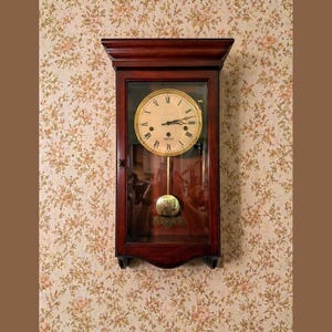 May include: A dark wood wall clock with a glass front, featuring a gold-colored clock face with black Roman numerals and a pendulum. The clock is mounted on a floral wallpaper background.
